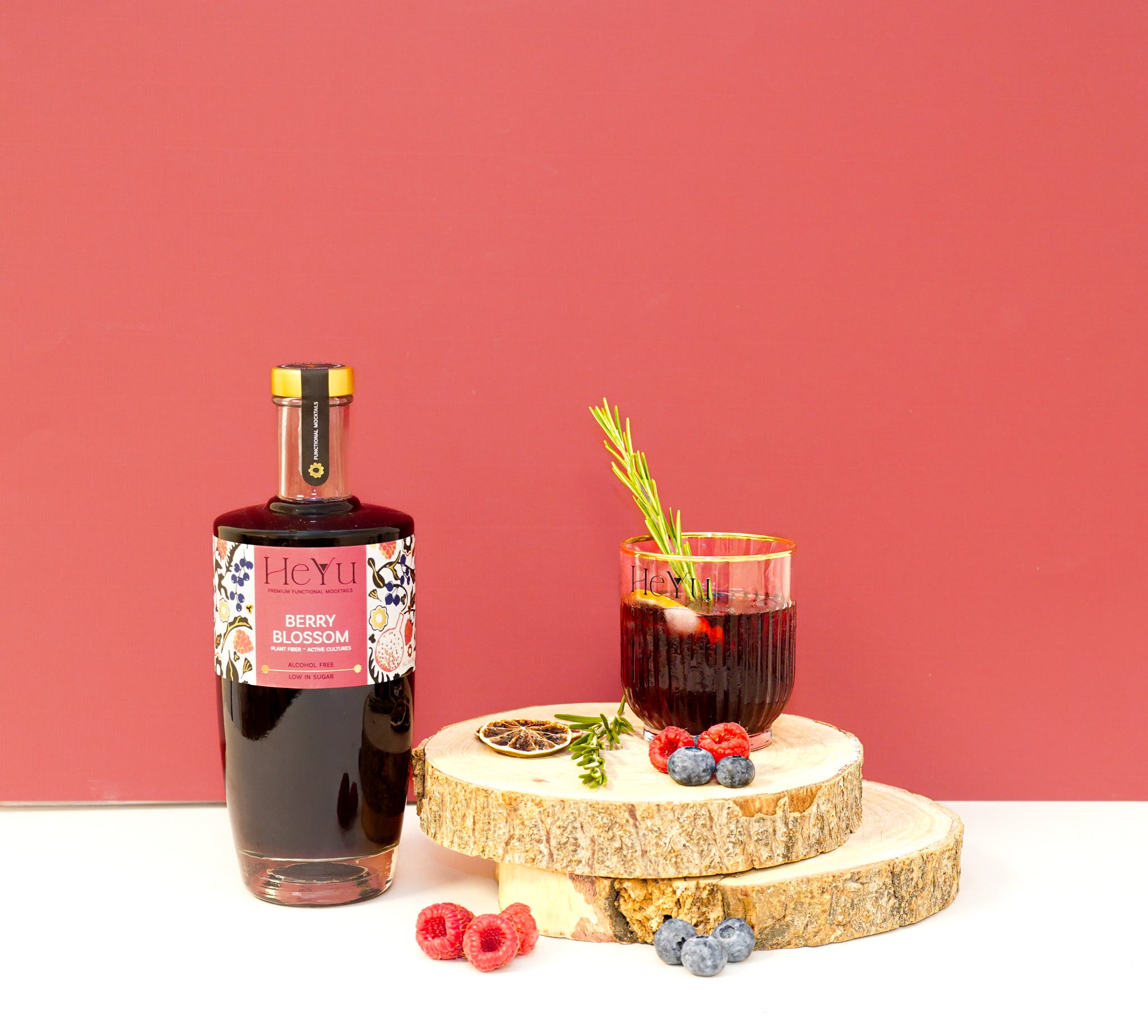 Bottle of HeYu Berry Blossom mocktail with a glass of cocktail on a wooden stand against a red background