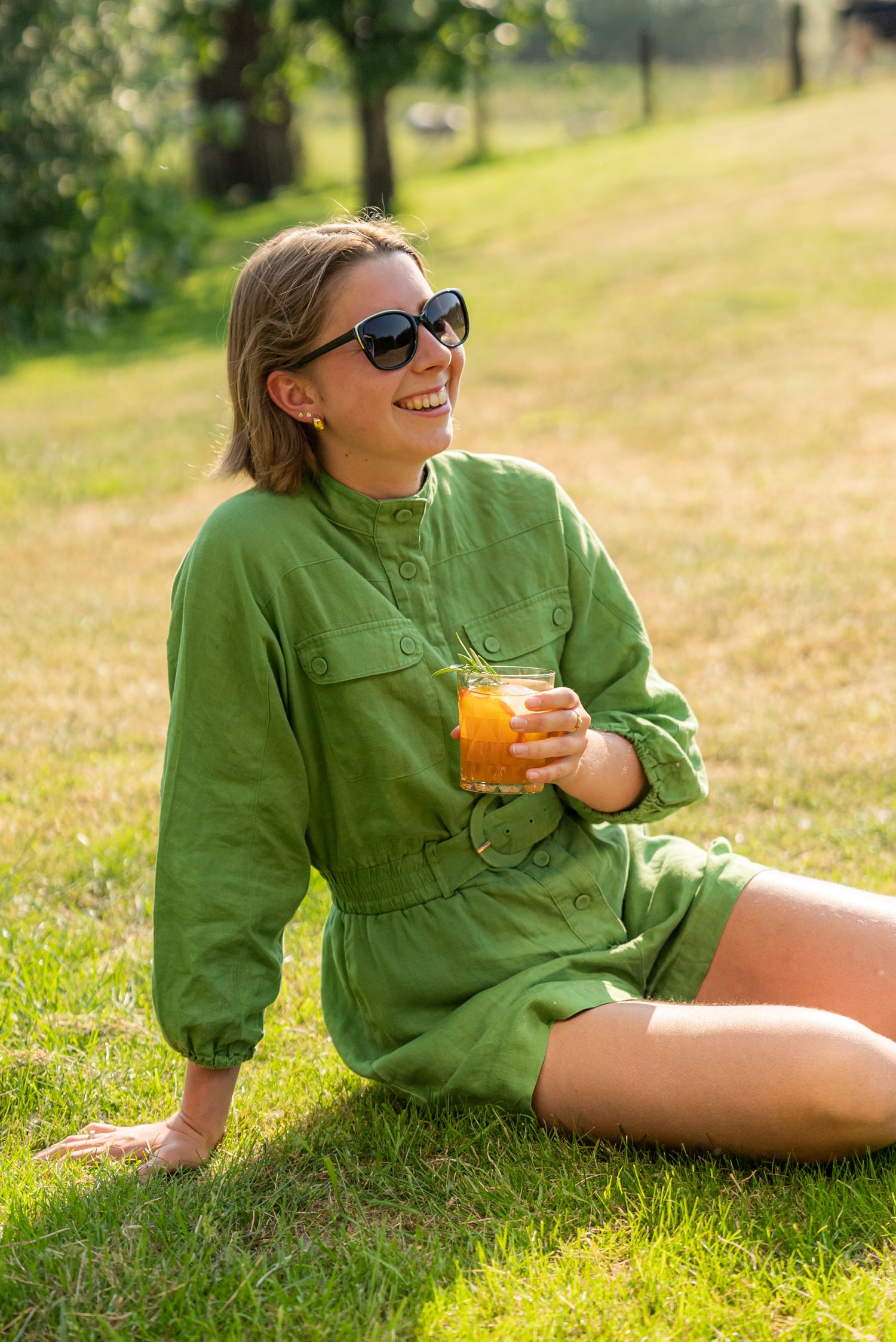 A woman in a green dress sitting on the grass, enjoying a glass of Heyu with a sprig of rosemary and ice.